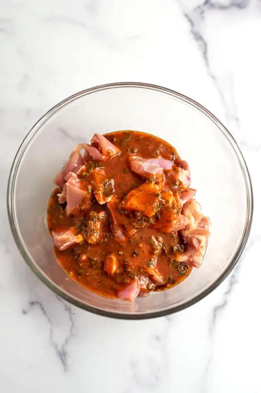 Raw chicken cubes in glass bowl with buffalo sauce poured over top.