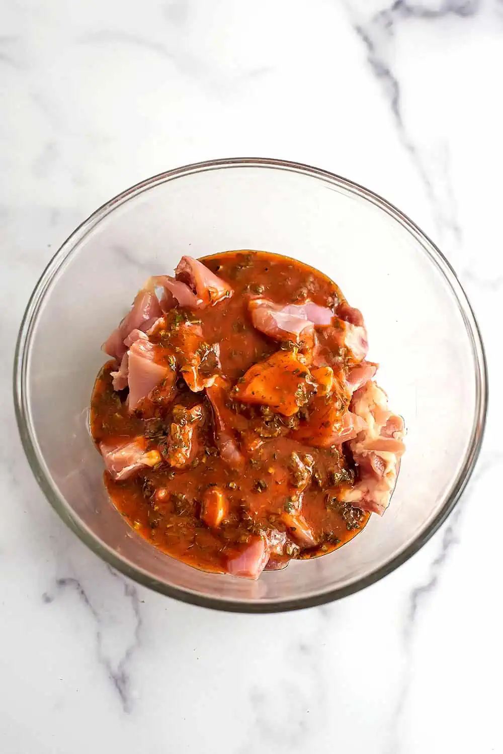 Raw chicken cubes in glass bowl with buffalo sauce poured over top.