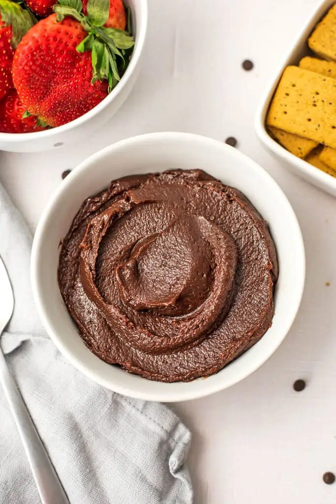 Thick chocolate tahini dip in a bowl with crackers and strawberries behind them.