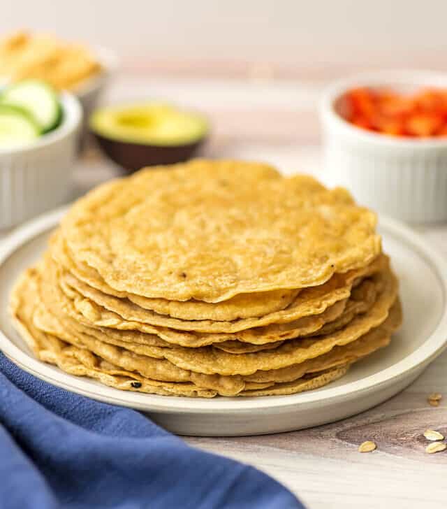 Oatmeal tortillas stacked on a plate with blue napkin and veggies in background.