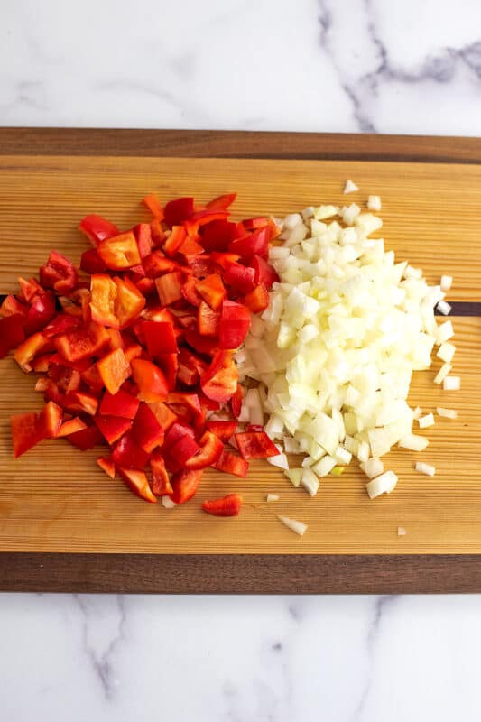 Chopped red bell peppers and onion on a wood cutting board.
