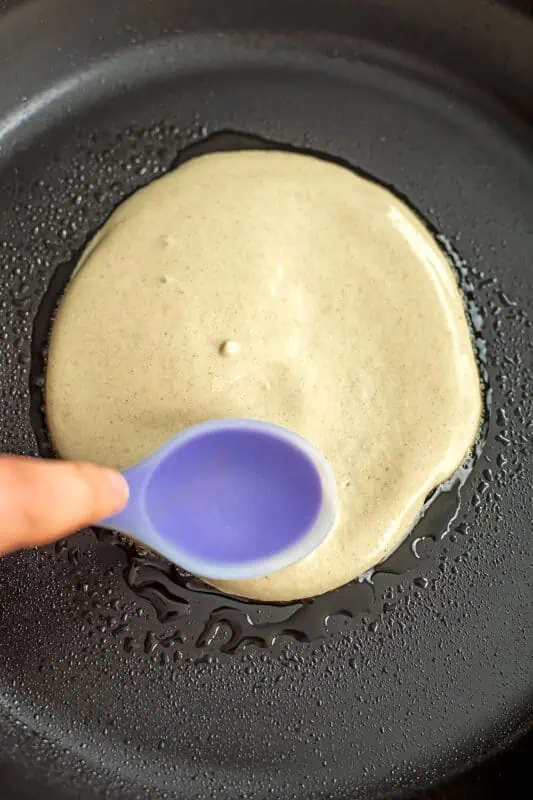Blue spoon spreading out oat flour batter in skillet.