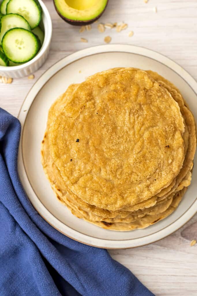 Oat flour tortillas stacked on a plate, bowl of cucumbers in background.