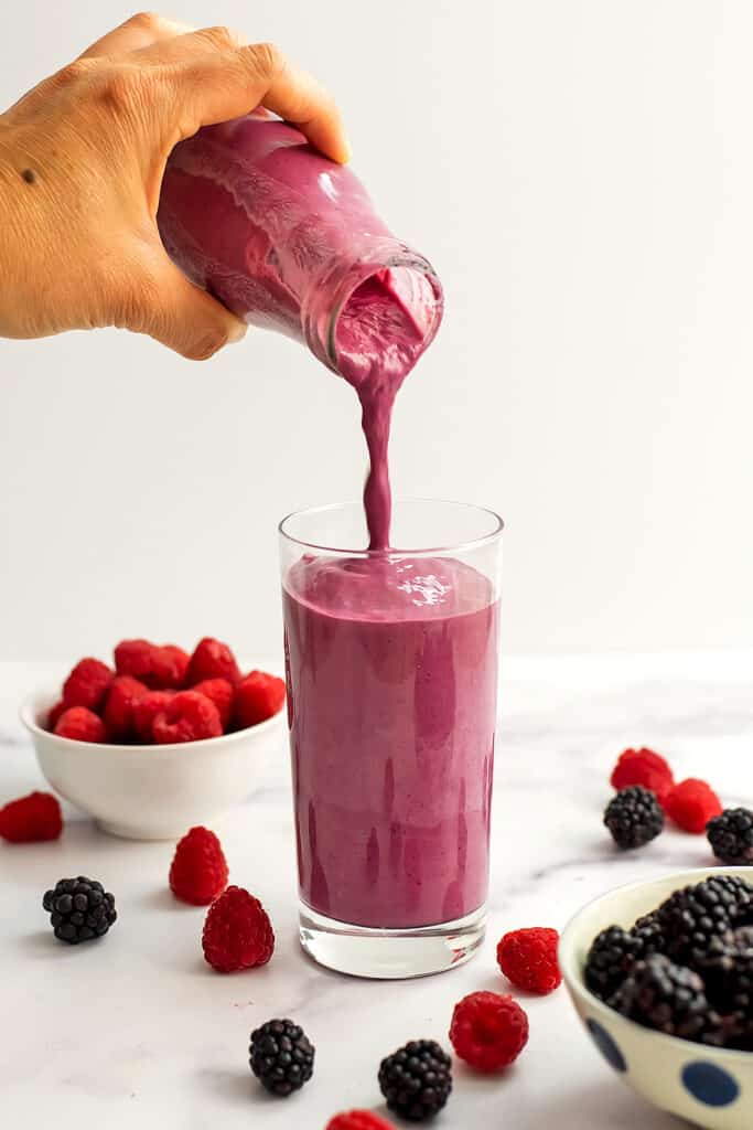 Blackberry Raspberry Smoothie being poured into a glass with blackberry and raspberry fruit piled high in bowls and laying on the table.