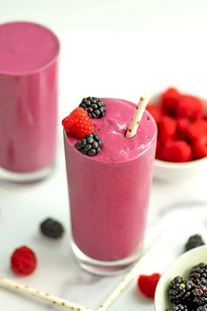 A blackberry raspberry smoothie in a glass with bright red raspberries in a bowl out of focus behind the smoothie.