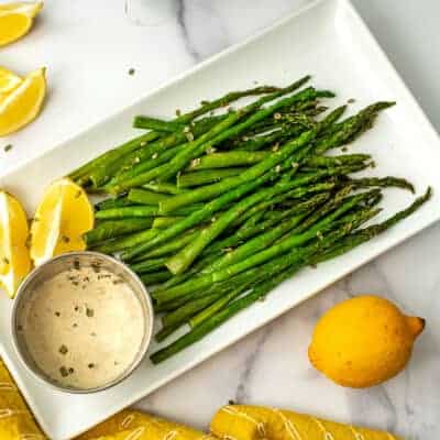 Cooked asparagus on a white plate with a ramekin filled with lemon tahini sauce.