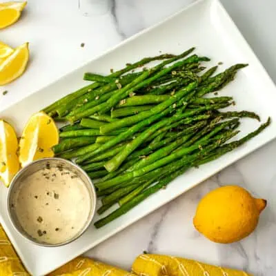 Cooked asparagus on a white plate with a ramekin filled with lemon tahini sauce.