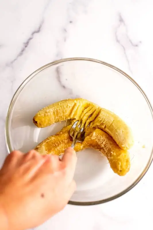hand mashing bananas in a glass bowl.