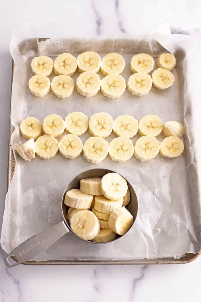 Frozen bananas slices on a baking sheet being placed a measuring cup.