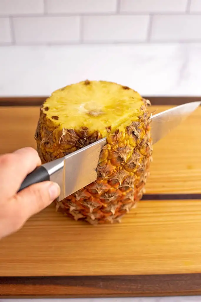 A whole pineapple on a cutting board being cut down the side removing it's skin.