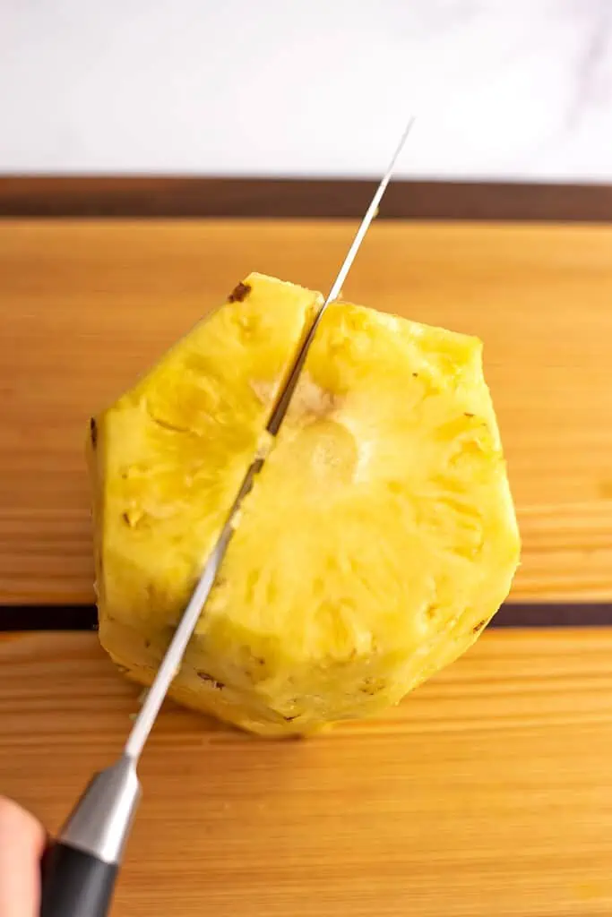 A whole pineapple on a cutting board with it's outer skin removed being cut in half.