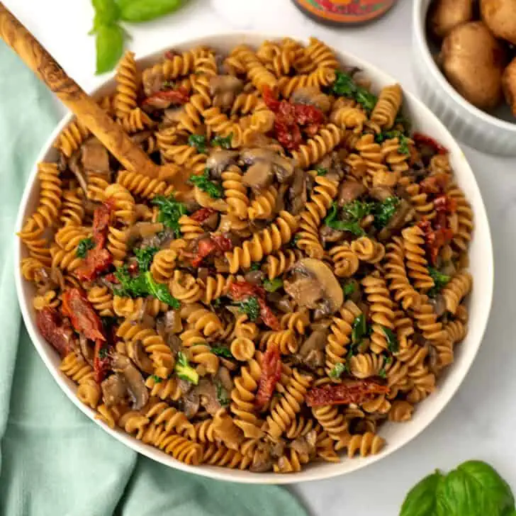 White bowl filled with mushroom sundried tomato pasta with wooden spoon.