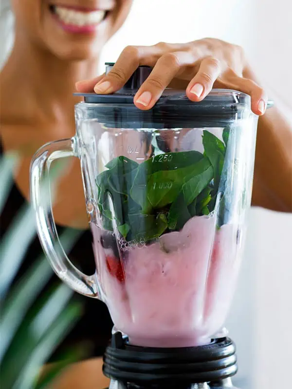A woman holding closed a blender in a kitchen. She is about to blend several ingredients into a smoothie in the blender.