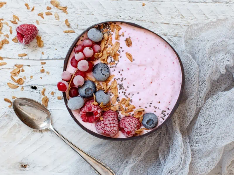 A pink smoothie in a bowl with raspberries, blueberries, granola and chia seeds on top.