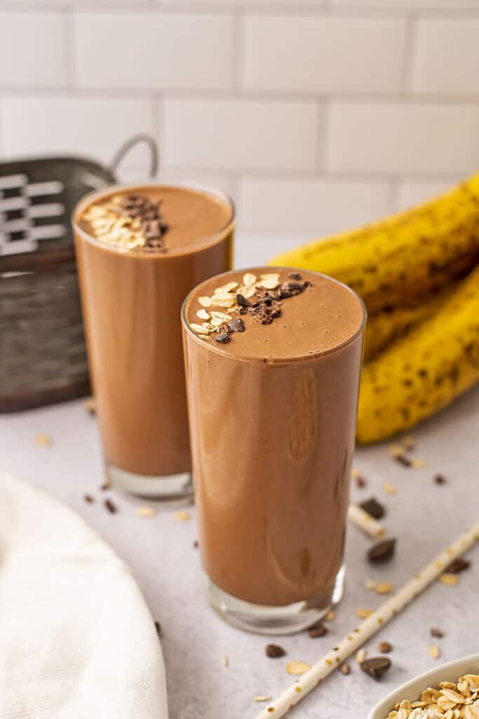 Chocolate banana oat smoothies sitting on as white counter top with oats and cacao nibs sprinkled on top.