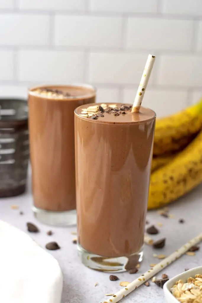 Chocolate oat smoothies with straws and toppings sitting on a white counter top with bananas in the background.