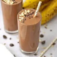 Oat Chocolate Smoothie with a straw on a white table with bananas in the background.