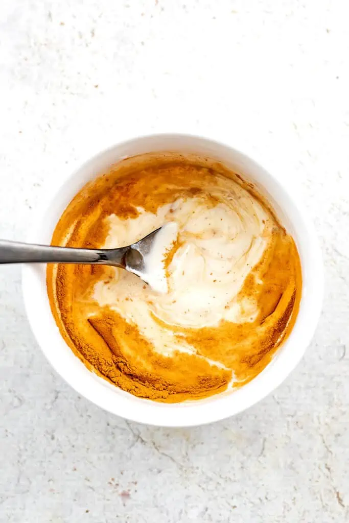 Spoon stirring cinnamon yogurt frosting ingredients in white bowl.