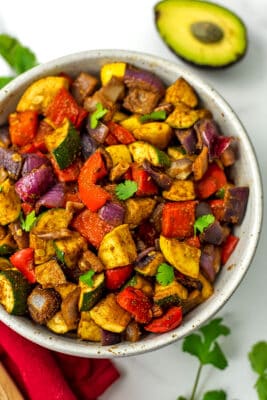 Mexican roasted vegetables in a white bowl with cut avocado.