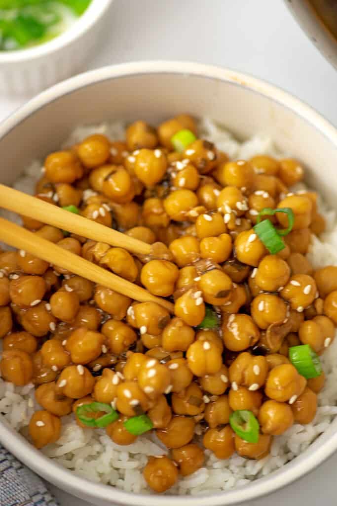 Wooden chopsticks picking up teriyaki chickpeas from a bowl.
