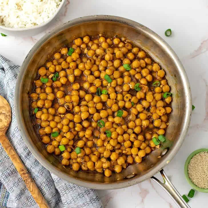 Teriyaki chickpeas in a skillet with a blue napkin and a spoon on the side.
