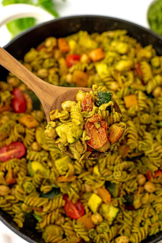Wooden spoon of pesto vegetable pasta over a skillet full of pasta.