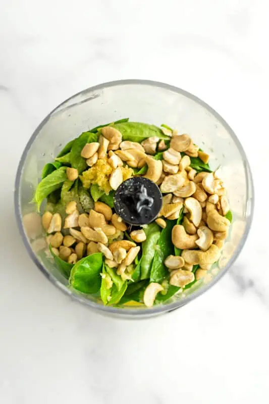 Cashews added to food processor with pesto ingredients.