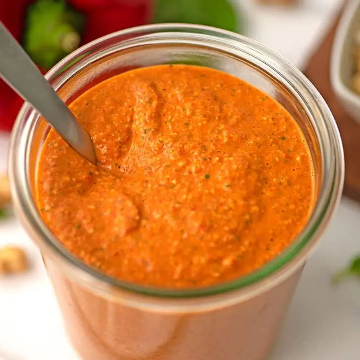 Large jar filled with red pepper pesto, spoon resting in jar.