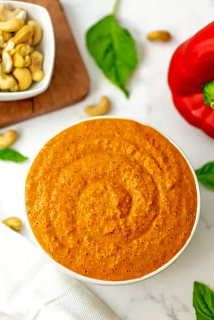 Red pepper pesto in a white bowl, peppers and cashews in background.