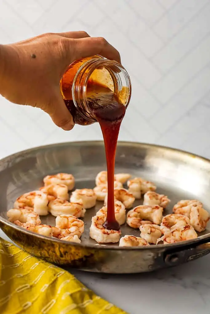 Honey sriracha sauce being poured over cooked shrimp in skillet.