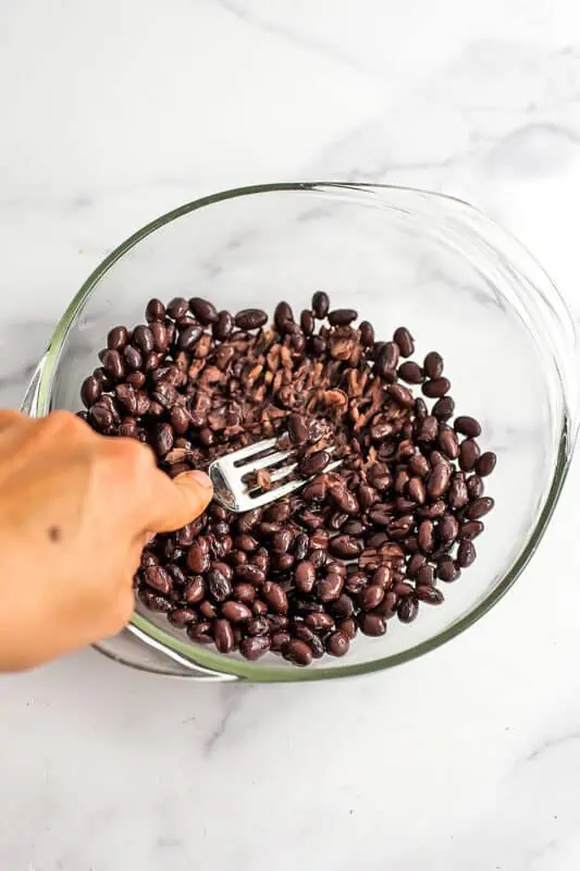 Fork mashing black beans in glass bowl.