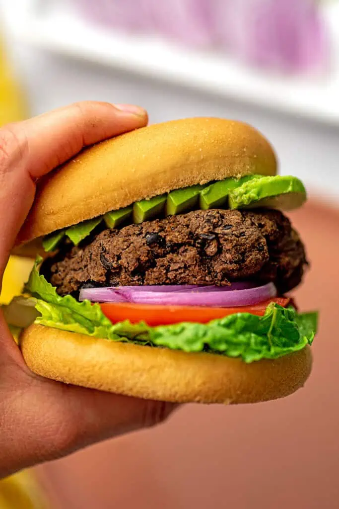 Hand holding black bean burger on a bun with lettuce, onion and tomato.