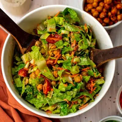 Salad servers resting in a bowl filled with spicy chickpea salad.