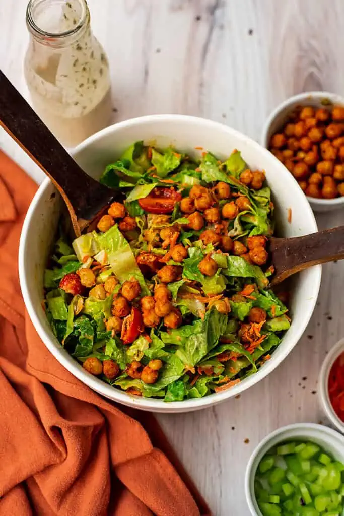 Buffalo chickpea salad in a white bowl with salad servers in the bowl.