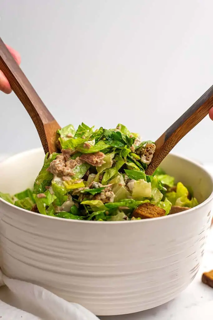 Wood serving spoons mixing up tuna caesar salad in white bowl.