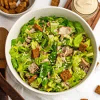 Caesar tuna salad in large white bowl with croutons and dressing in background.