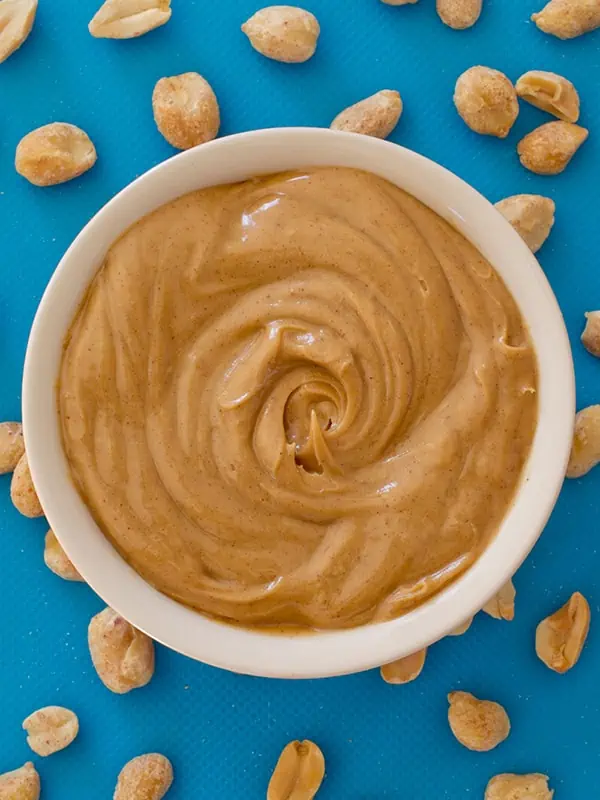 Creamy peanut butter in a bowl with peanuts scattered around it on a blue surface.