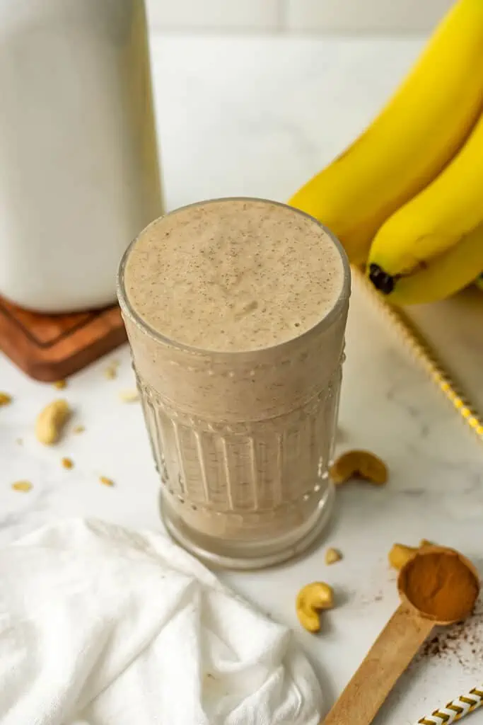 Banana bread smoothie in a glass with bananas and milk in the background.