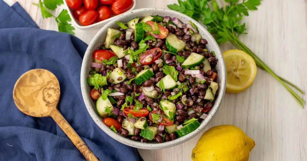 Mediterranean black bean salad in a white bowl next to a wooden spoon and blue napkin.