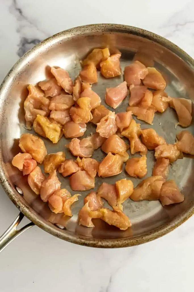 Raw chicken cubes being cooked in a stainless steel skillet.