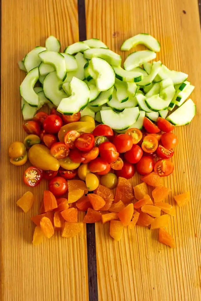 Cut up cucumber, tomatoes and dried apricots on a wooden cutting board.