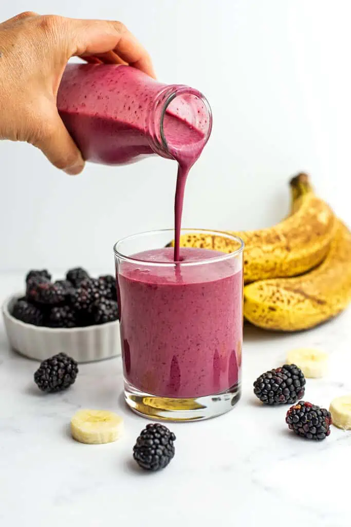 Blackberry banana smoothie being poured into a glass with pieces of blackberry and cut bananas laying around it on a white table.