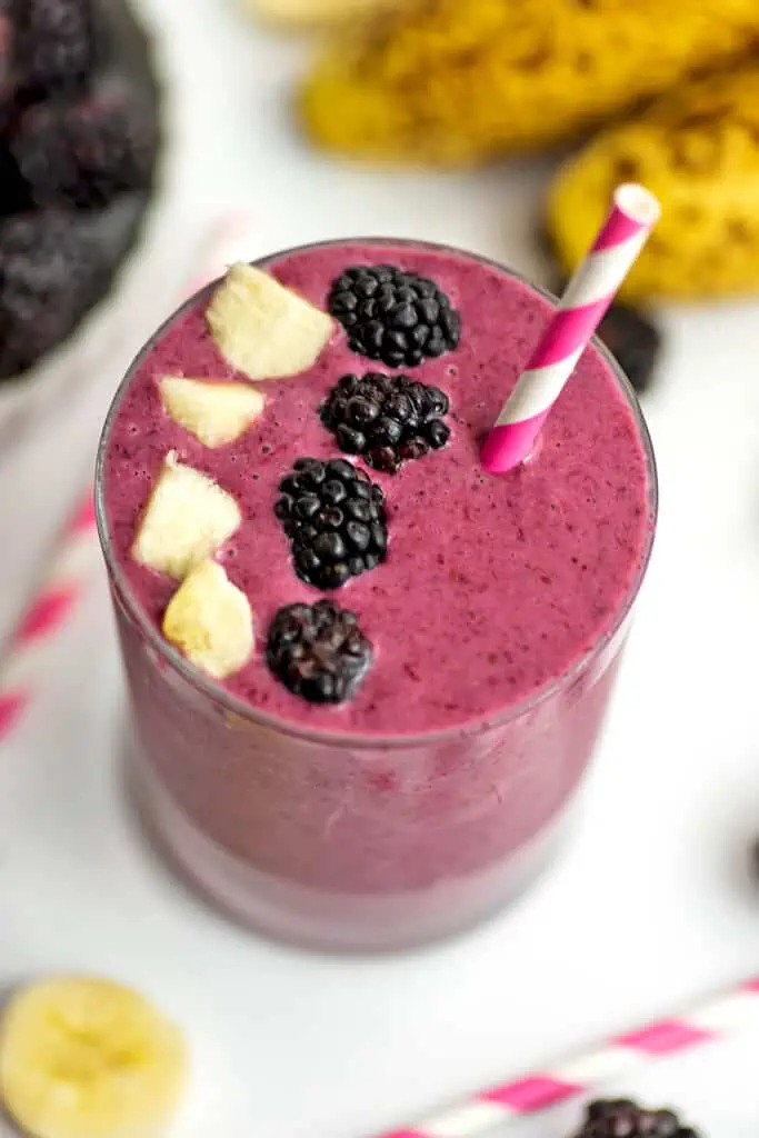 A blackberry banana smoothie in a small glass with blackberries and pieces of banana on top with a straw.