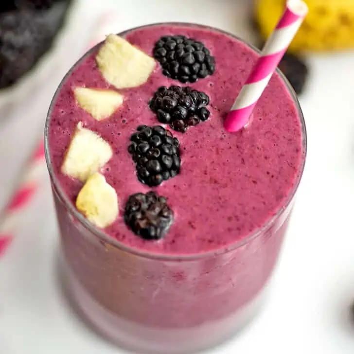 A blackberry banana smoothie in a small glass with blackberries and pieces of banana on top with a straw.