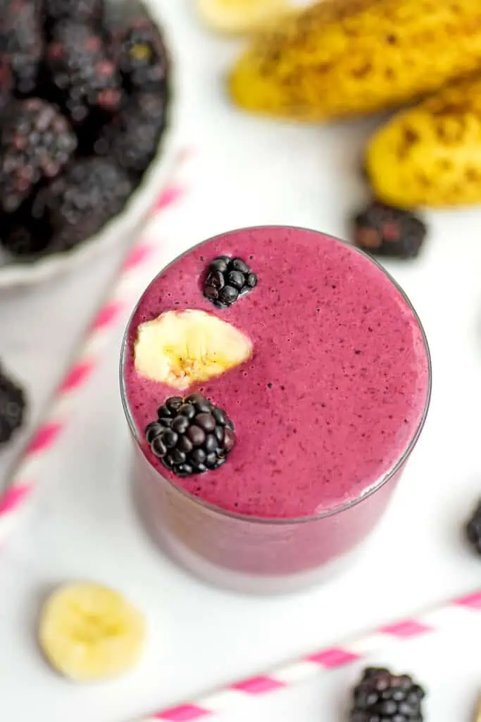 Blackberry banana smoothie with whole blackberries and cut bananas in a glass. Blackberries in a bowl are out of focus behind the smoothie. 