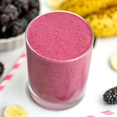 A small cup full of Blackberry Banana Smoothie sitting on a white table with fruit around it.