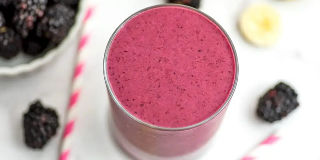 A close overhead view of a blackberry banana smoothie with paper straws and whole blackberries around the smoothie.