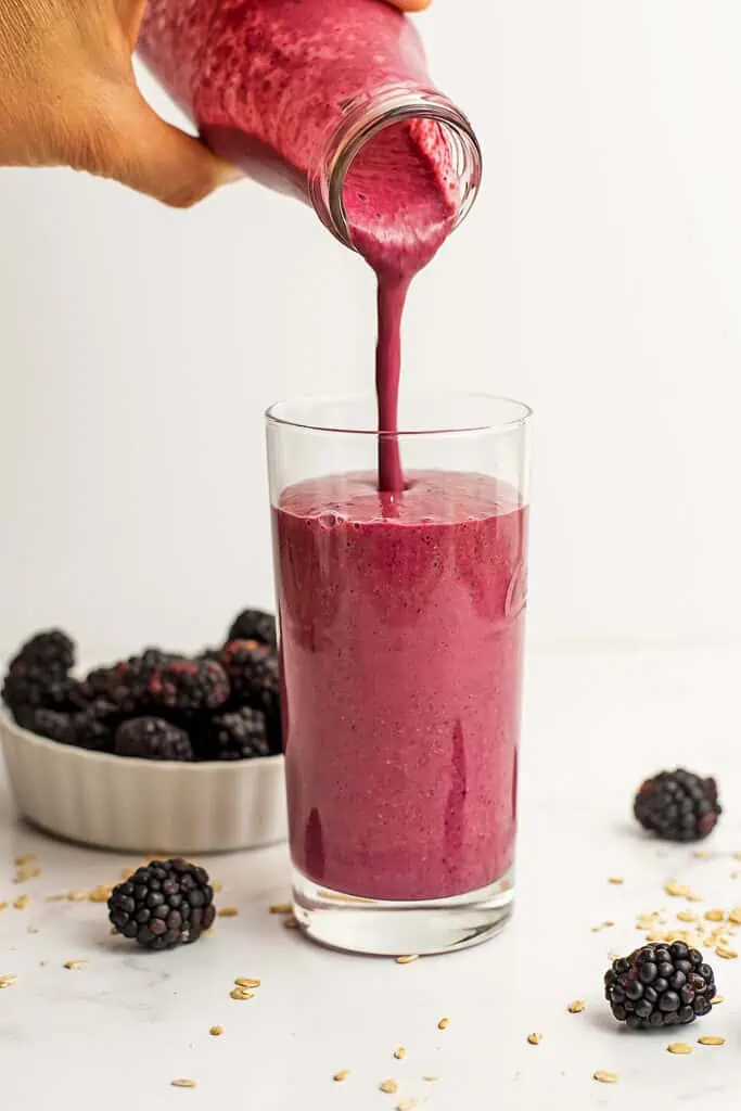 Pouring blackberry oatmeal smoothie into a glass with blackberries in a white ramekin in the background.