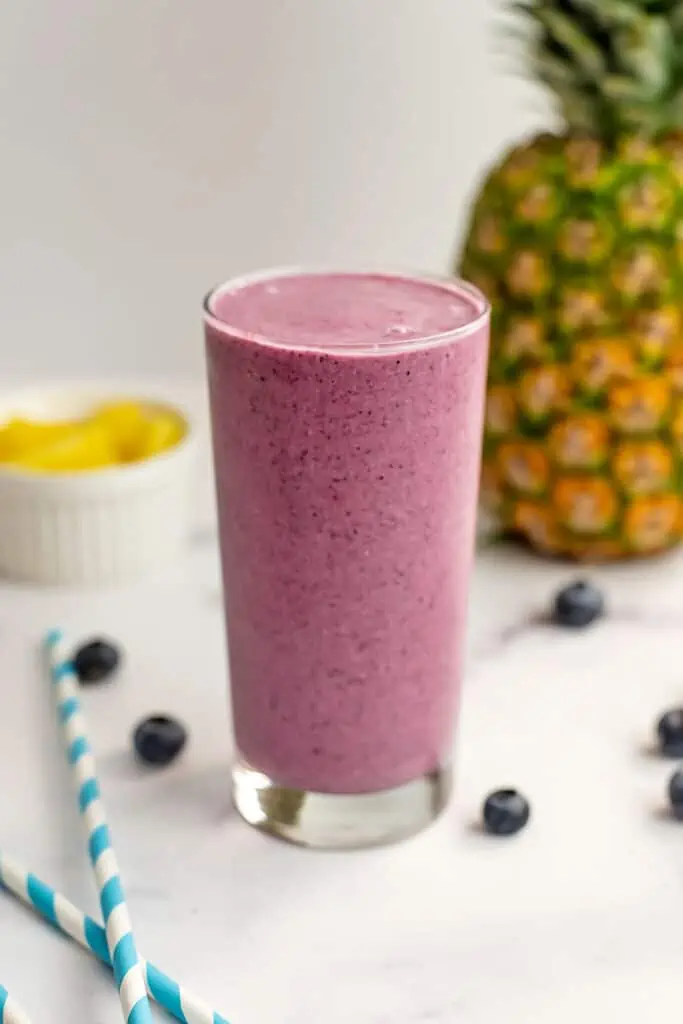 A pineapple blueberry smoothie sitting on a white table. A whole pineapple and pineapple chunks in a white ramekin are just out of focus behind the smoothie.