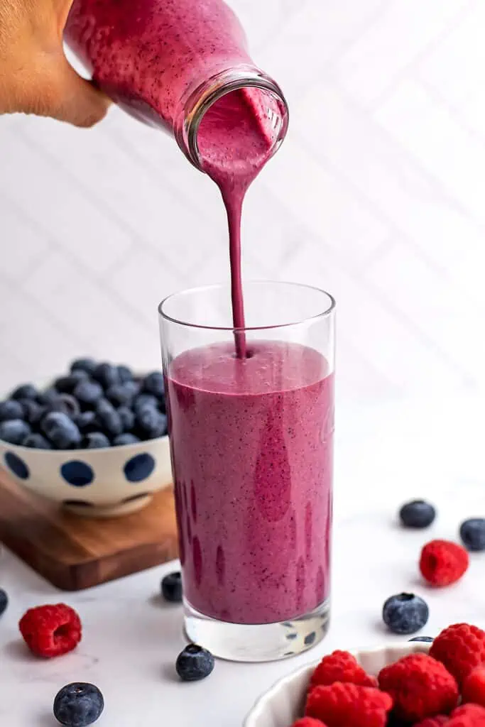 A raspberry blueberry smoothie being poured into a tall glass on a white table. 